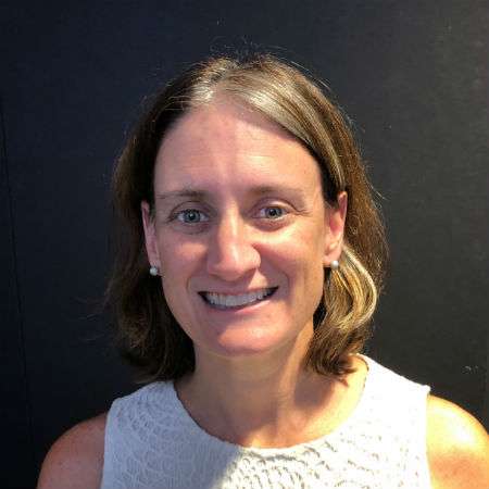 A woman with shoulder-length brown hair, wearing a sleeveless white top, smiles at the camera against a plain dark background.