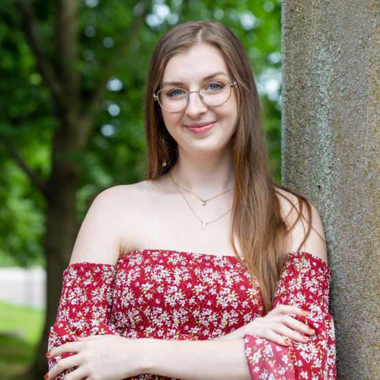 A young woman with long brown hair and glasses stands outdoors by a stone pillar, wearing a red floral off-shoulder dress and smiling at the camera. Trees are visible in the background.