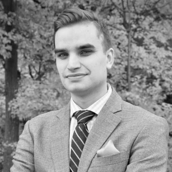 A man in a suit and tie stands outdoors with arms crossed, posing in front of trees. The image is in black and white.