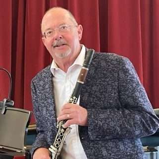 A man in a patterned jacket holds a clarinet, standing in front of red curtains with music equipment nearby.