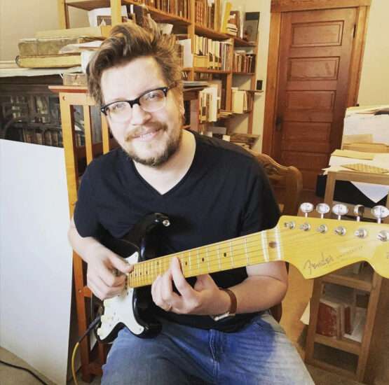 A man with glasses and a beard sits indoors, playing a black and white Fender electric guitar, surrounded by bookshelves.