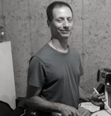 A man in a t-shirt stands indoors, holding drumsticks, next to a table with papers and audio equipment. The background appears to be a concrete or unfinished wall.