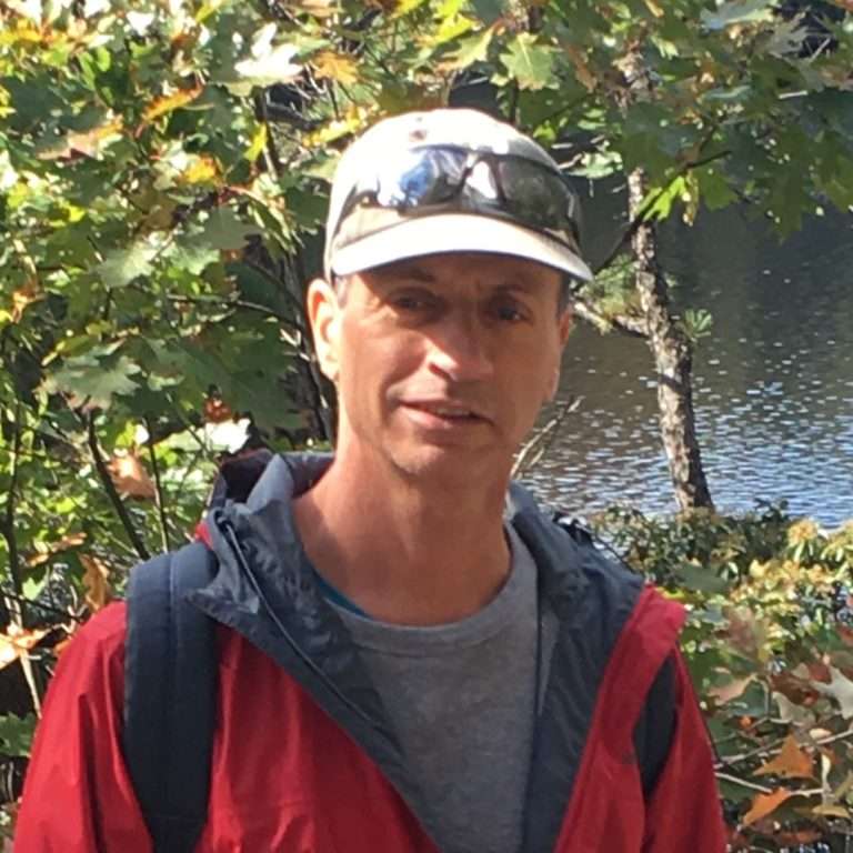 Man wearing a red jacket, gray shirt, and cap with sunglasses, standing outdoors near trees and a body of water.