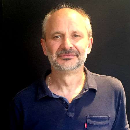 A middle-aged man with short, graying hair and a beard is standing against a plain dark background, wearing a navy blue collared shirt.