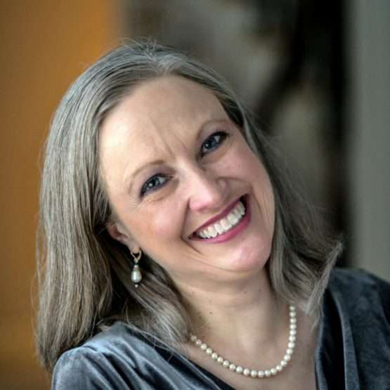 Smiling woman with straight gray hair, wearing a pearl necklace, pearl earrings, and a gray top, photographed indoors with a blurred background.