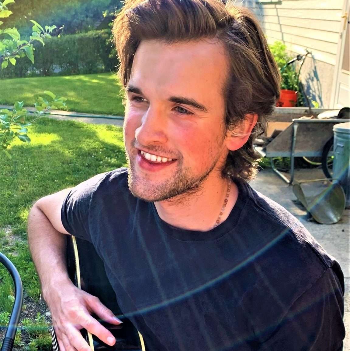 A man with medium-length hair sits outside in sunlight, playing an acoustic guitar and smiling. There is grass and a wheelbarrow in the background.
