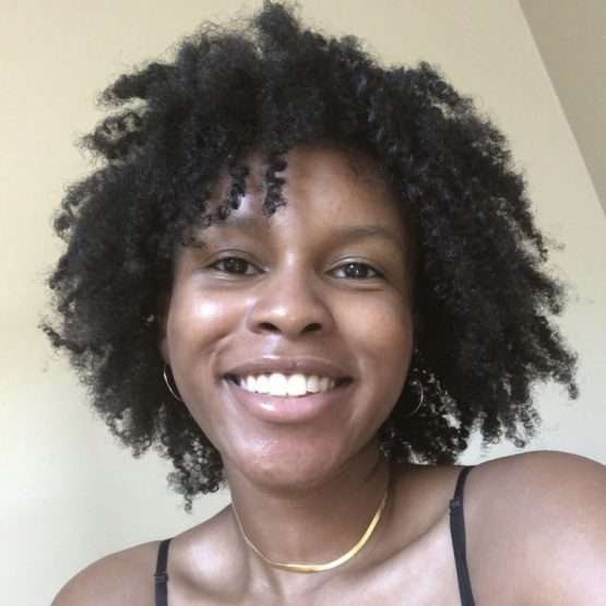 A person with curly hair smiles at the camera, wearing hoop earrings, a thin gold necklace, and a black strap top, against a plain beige background.