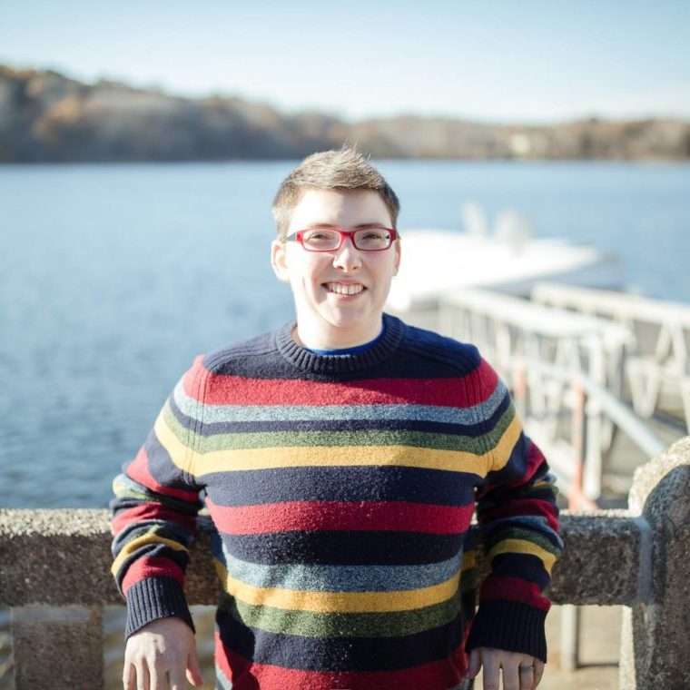 Person wearing glasses and a striped sweater stands by a stone railing in front of a lake on a sunny day.