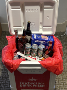 A white Igloo cooler lined with a red bag is filled with various alcoholic drinks, including cans, bottles of liquor, and a wine bottle. The cooler lid is open and “Budweiser Drink Wiser” is printed on the front.