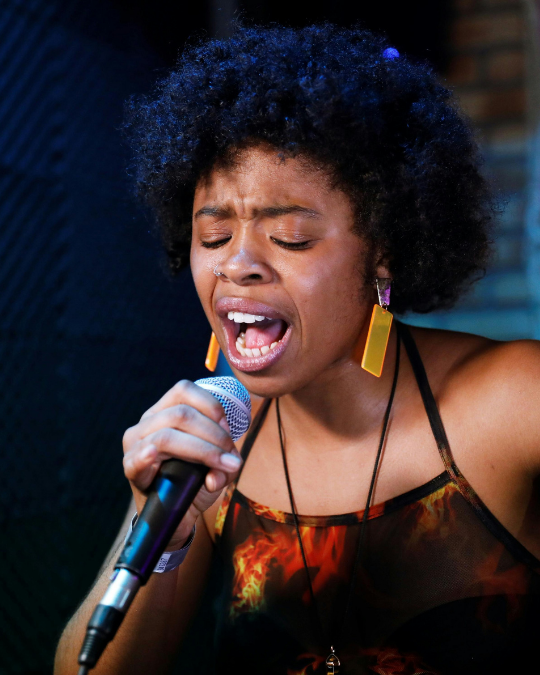 A woman with curly hair passionately sings into a microphone. Wearing large orange earrings and a colorful sleeveless top, she is deeply immersed in her performance.