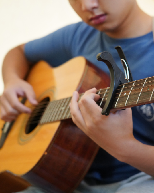 A person in a blue shirt plays an acoustic guitar, focusing on the strings and fretboard with a capo attached to the neck. The slightly blurred image hints at participation in group music classes, and their full face is not visible.