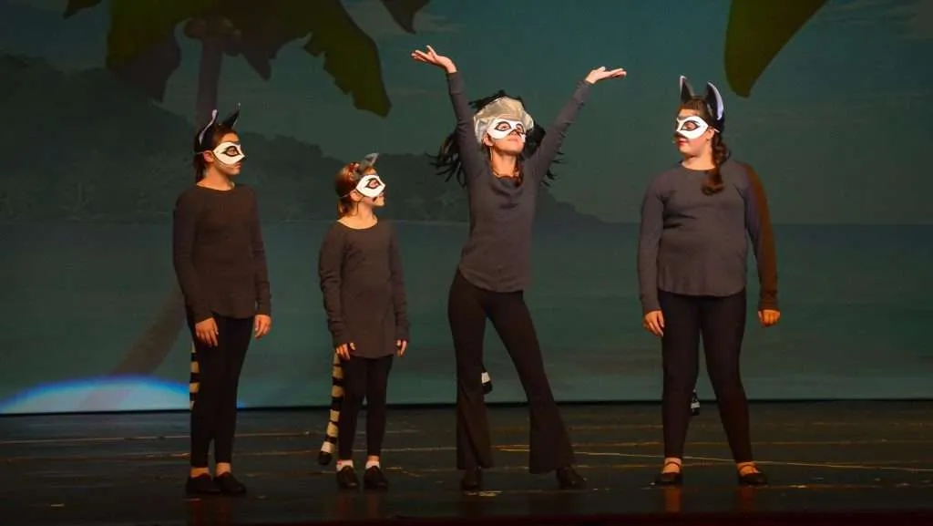 Four children in lemur costumes and face paint stand on stage, with one raising their arms while the others look on, against a backdrop of a tropical scene.