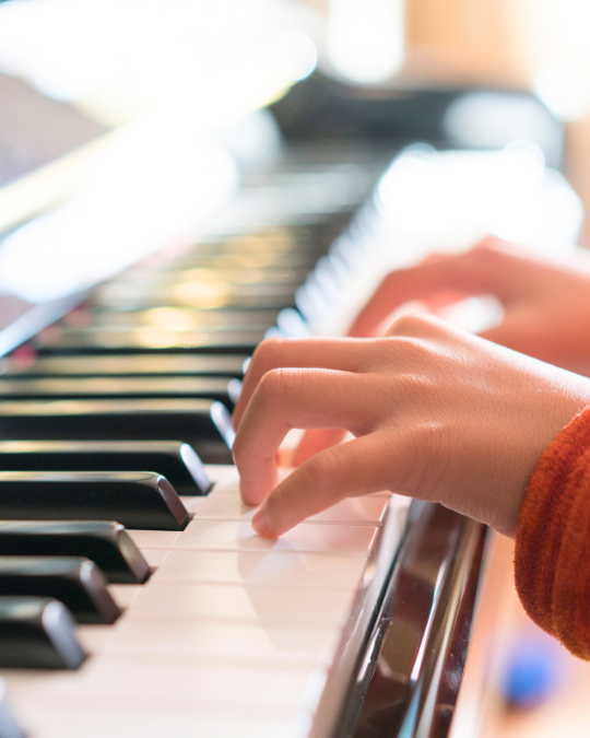A close-up of a person’s hands playing piano keys, softly lit by natural light. The focus on the fingers suggests concentration and musical practice, often developed in music programs or group music classes.