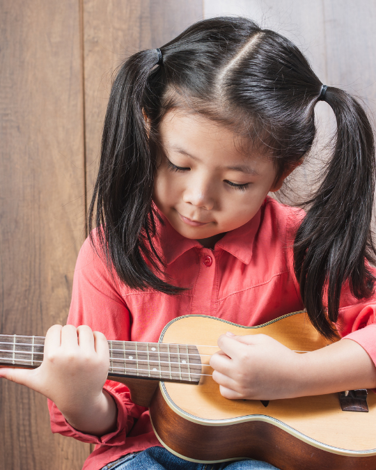 A young girl with black pigtails and a pink shirt is playing a small acoustic guitar or ukulele. She is looking down at the instrument, focused on her fingers as she strums, practicing for her group music classes.