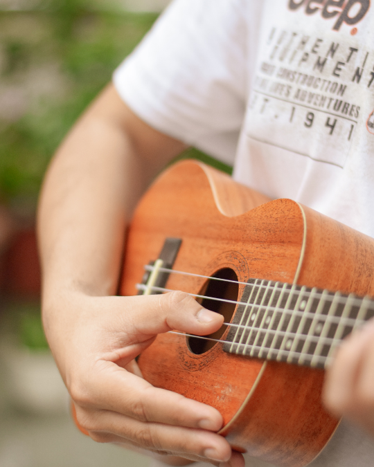 A person in a white t-shirt is playing a small wooden ukulele, focusing on their hand strumming the strings—perfect inspiration for Group Music Classes or Music Programs. The blurred background features green plants.