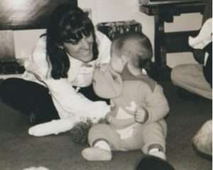 A woman smiles at a baby in a onesie, both enjoying a candid moment indoors. This black-and-white photo captures the warmth found in Apple Tree Arts’ community arts programs.
