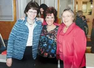 Three smiling women, Kathy, Jan and Donna in the early 2000s, stand close together indoors, representing Apple Tree Arts. The woman on the left wears glasses and a blue jacket; the center has spiky hair and a colorful top; the right, in a red jacket, shares their passion for arts education and community music.