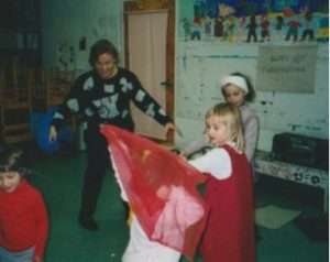 A group of children play with a red scarf in a classroom while an adult watches. The space, filled with children’s artwork and a “Happy Thanksgiving” sign, reflects the lively spirit of community arts at Apple Tree Arts.