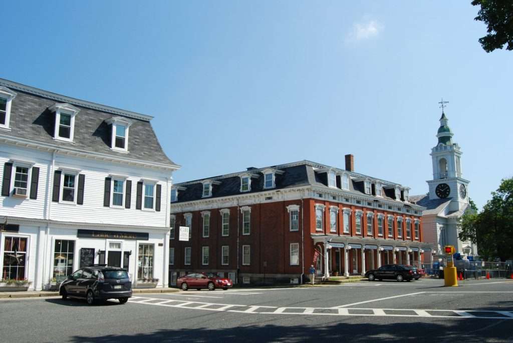 A sunny day in a quaint town center with historic buildings, including a white shop, a red brick building, and a church with a clock tower—home to Apple Tree Arts, offering music classes and arts education—cars parked along the street beneath a clear blue sky.