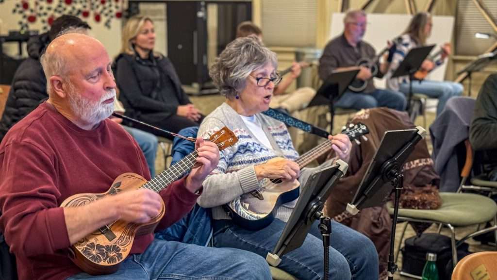 Group of adults seated indoors, playing ukuleles and reading music from stands during a ukulele workshop.
