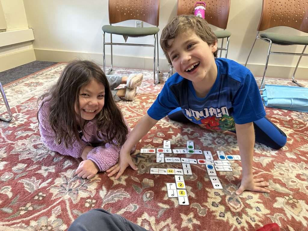 Two smiling children sit on a patterned carpet, playing a music tile game together. The boy wears a blue shirt and the girl a purple sweater, chatting happily during an early childhood program. Chairs are visible in the background.