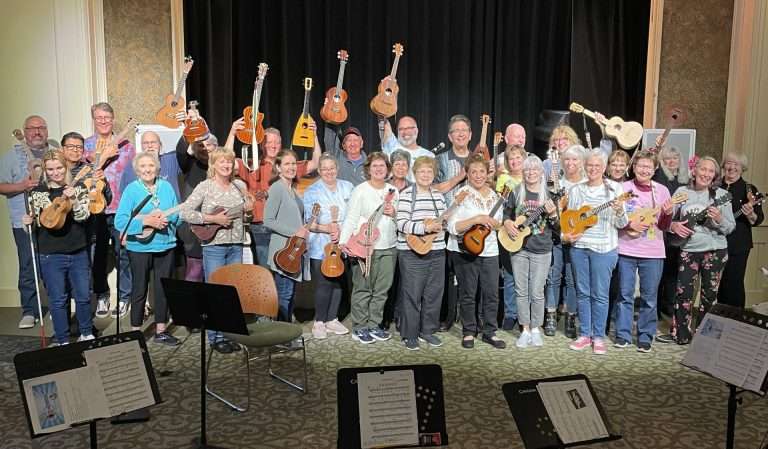 A large group of people from Apple Tree Arts stand on a stage holding ukuleles, smiling at the camera. Some raise their instruments in the air. Music stands with sheets are visible in front; a black curtain is in the background.