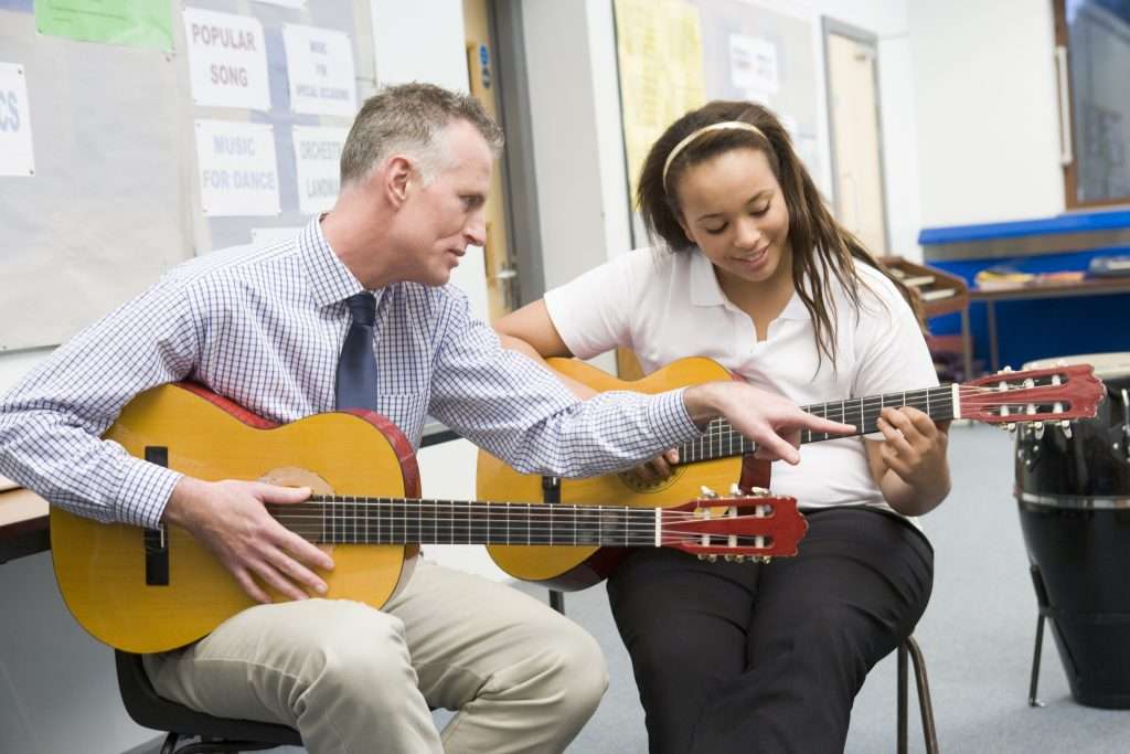A man is teaching a young woman to play guitar in a classroom. Both are holding acoustic guitars; the man is pointing at the woman’s fingers on the fretboard. The setting includes music-themed posters on the wall.