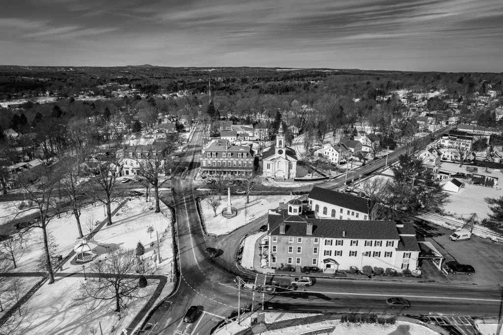 A black and white aerial view of a small town center in winter, featuring Apple Tree Arts among the prominent buildings in Grafton Common, with snow-covered rooftops and trees, and roads converging at an intersection.