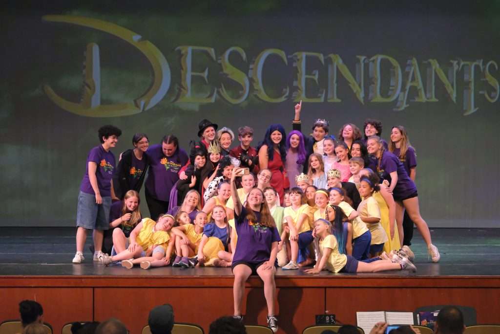 A large group of children and teens pose and smile on stage in front of a screen that says “Descendants.” Some wear purple shirts and others wear yellow; a few are in costume. The stage is set for a theater performance.