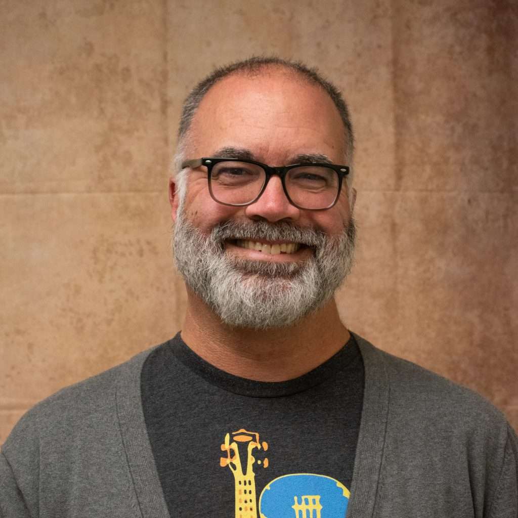A smiling man with glasses and a gray beard, wearing a dark t-shirt with a yellow graphic and a gray cardigan, stands in front of a plain, beige background.