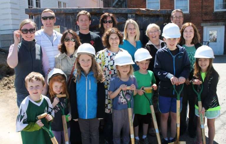 A group of adults and children pose outdoors at a construction site. The children in front hold shovels and wear white hard hats, while the adults behind them smile at the camera. A large metal dumpster is in the background.
