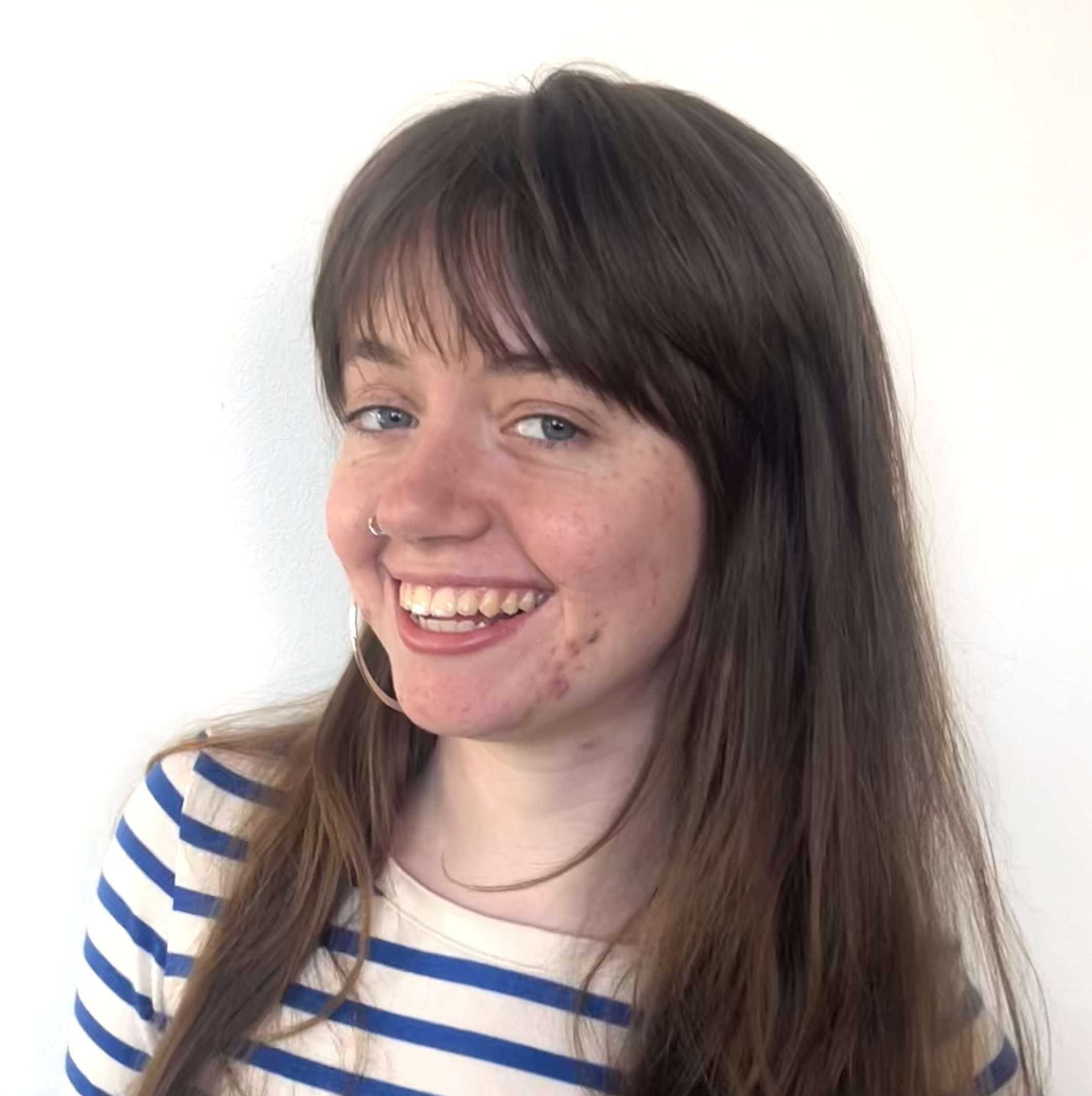 Young woman with long brown hair, wearing a white shirt with blue stripes, smiling in front of a plain white background.