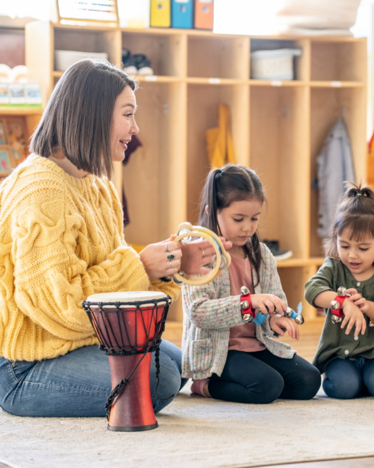 An adult and two young children sit on the floor playing musical instruments, including a drum, tambourine, and wrist bells, in a classroom setting.