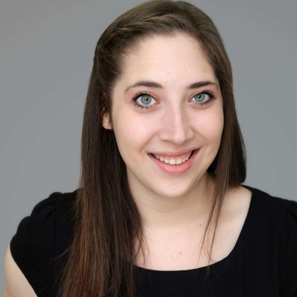 A woman with long brown hair and blue eyes, wearing a black top, smiles at the camera against a plain gray background.