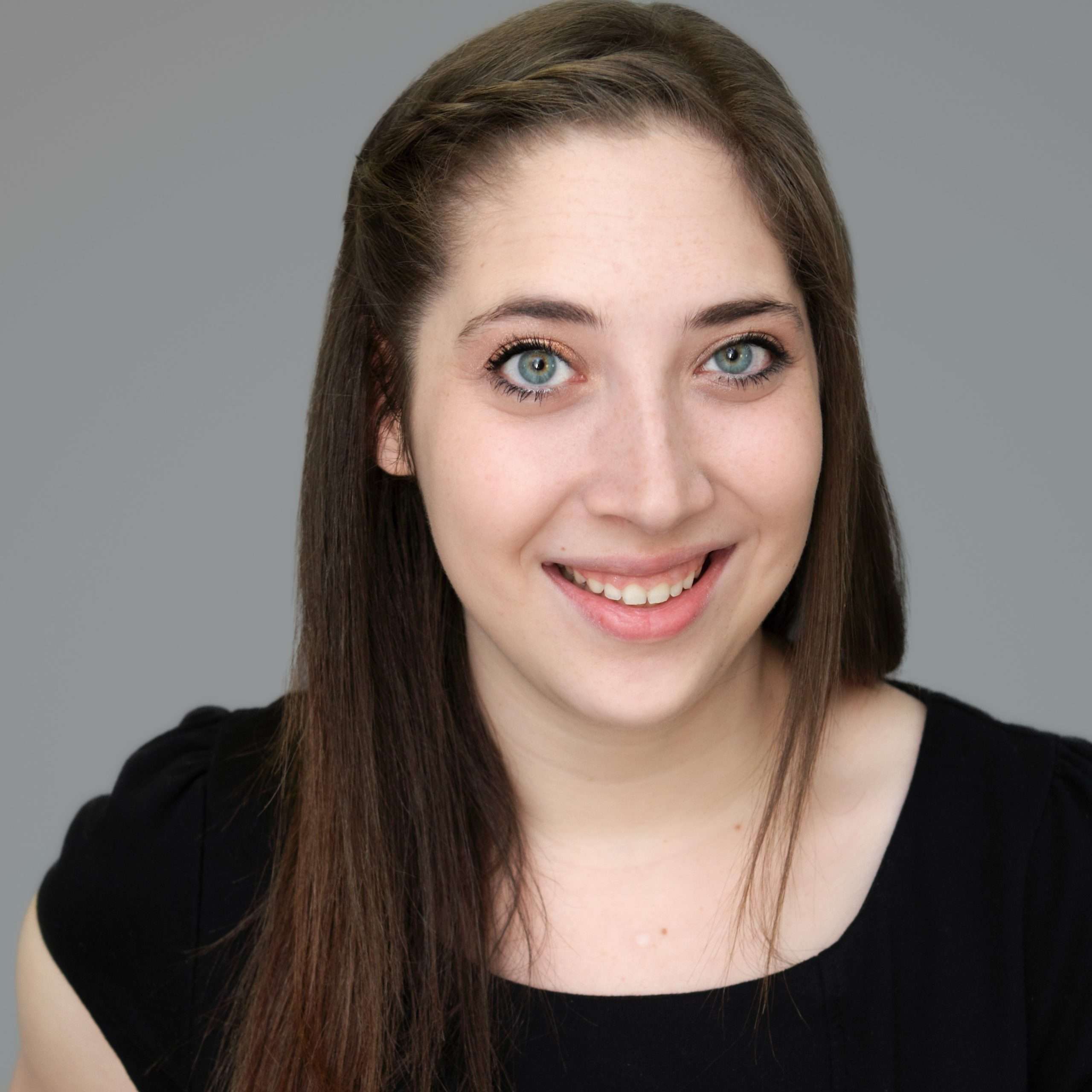 A woman with long brown hair and blue eyes, wearing a black top, smiles at the camera against a plain gray background.