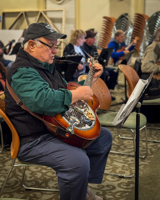 Man wearing a cap and glasses plays a guitar while seated, reading sheet music; several other musicians are visible in the background.