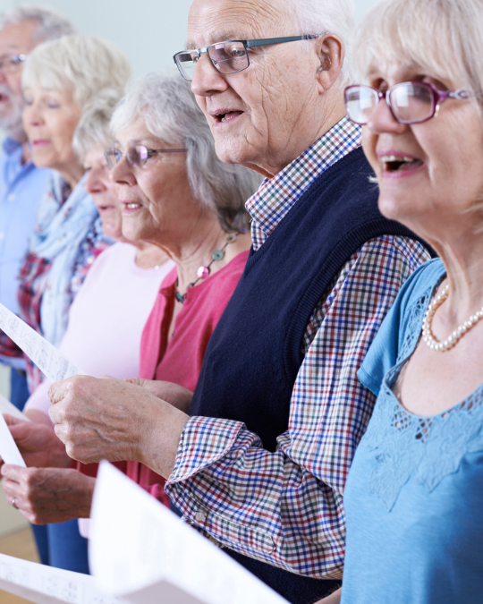 A group of older adults standing in a row, holding sheets of paper and singing together.
