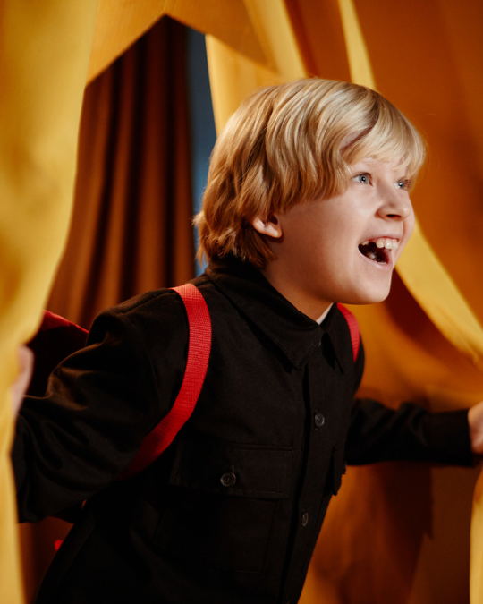 A smiling child wearing a backpack pulls back yellow curtains