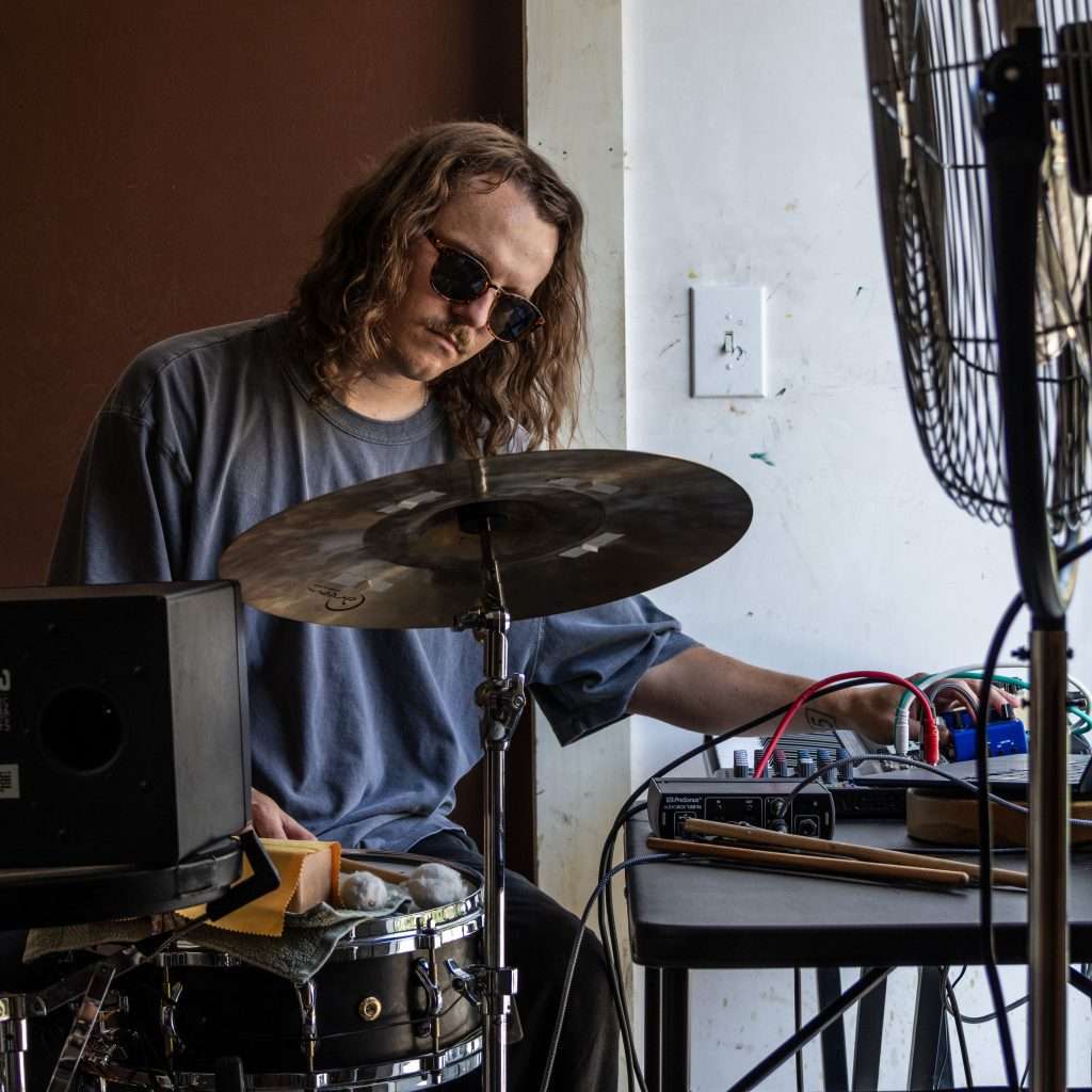 A person with long wavy hair and sunglasses plays drums, looking down with focus. They wear a gray shirt and are in a room with brown and white walls.