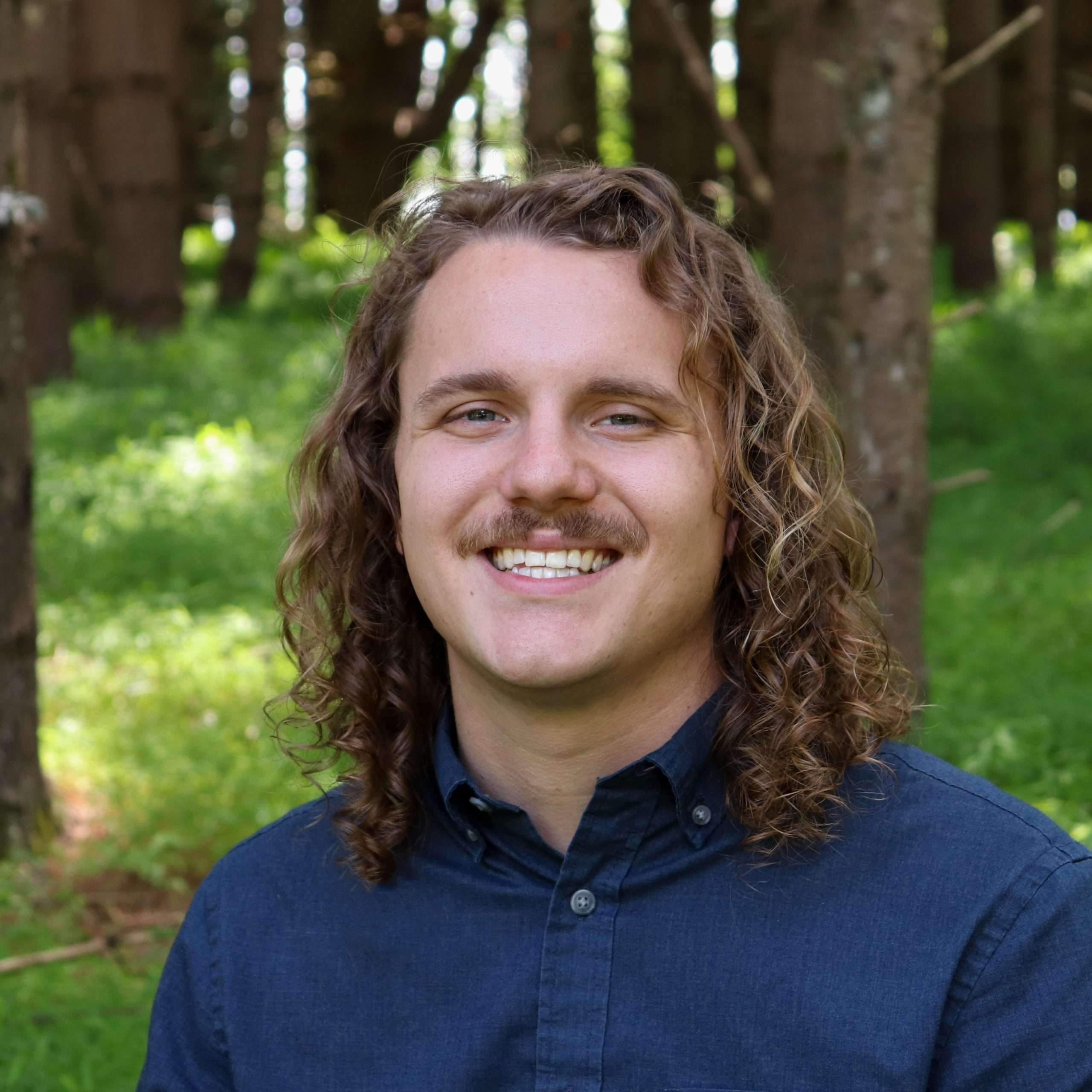 A person with long curly hair and a mustache, wearing a dark button-up shirt, smiles while standing in a forested area with green grass and tall trees.